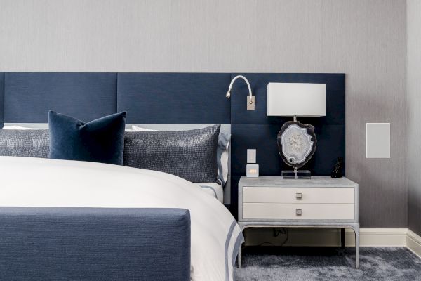 The image shows a modern bedroom with a navy blue headboard, pillows, a bedside table, and a lamp. It has a sleek, minimalist design.