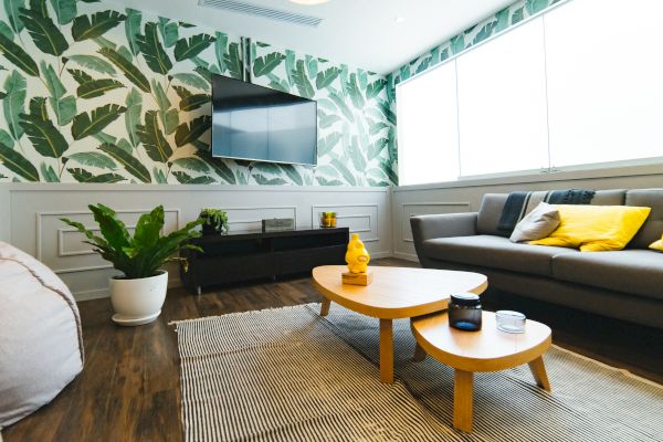The image features a modern living room with leafy wallpaper, a TV, a cozy couch, and stylish coffee table decor.
