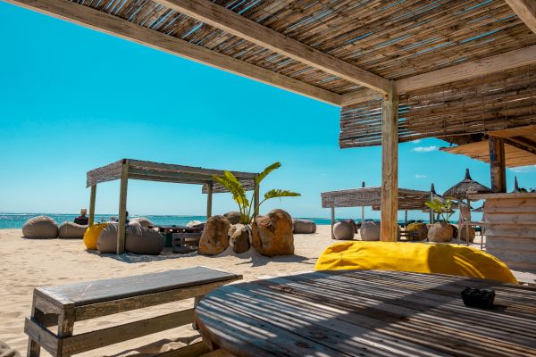 The image shows a beachside setting with wooden structures, lounge areas, and a clear blue sky. It looks inviting and relaxing.