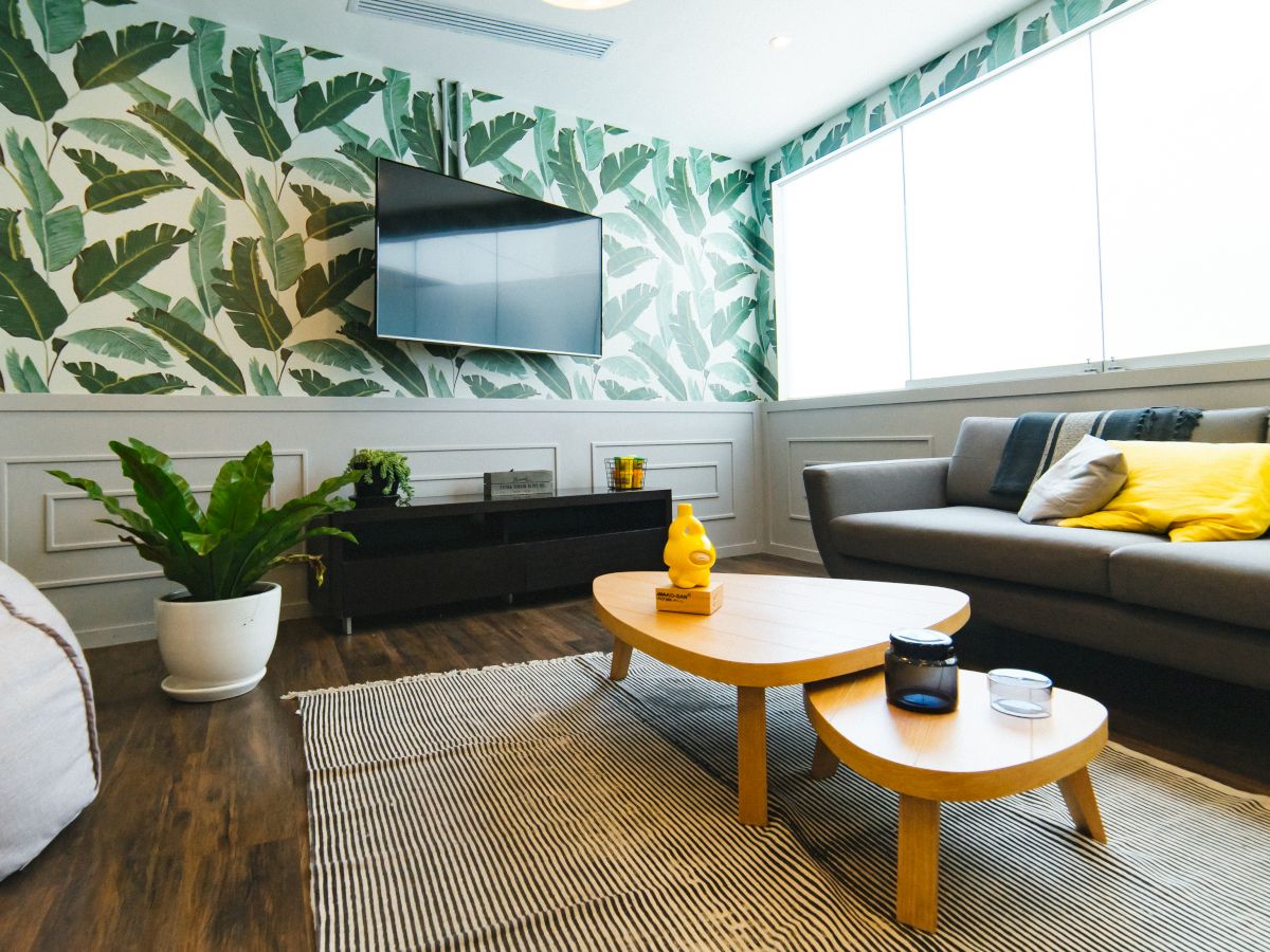 The image features a modern living room with leafy wallpaper, a TV, a dark sofa, yellow cushions, and a coffee table.
