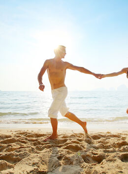 A couple joyfully runs along a beach, hand in hand, with sunlight glistening on the water behind them. It's a moment of happiness.