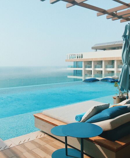 The image shows a luxurious pool area with lounge seating, a vibrant blue pool, and a serene ocean view in the background.
