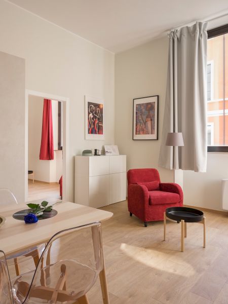 The image shows a modern, bright living space with a kitchen, dining table, and cozy seating area featuring red accents.