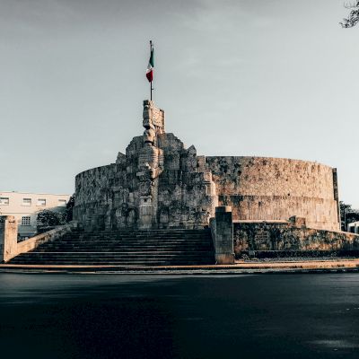 The image features a prominent historical monument with a circular base and a flagpole atop, surrounded by trees and a roadway.