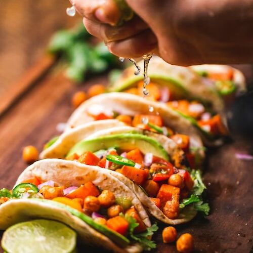 A hand squeezes lime juice over colorful tacos filled with vegetables, served on a wooden board with lime wedges.