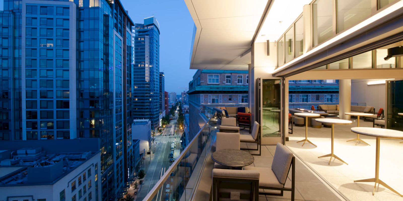 The image shows a sleek balcony overlooking a cityscape at dusk, with modern furniture and illuminated buildings in view.