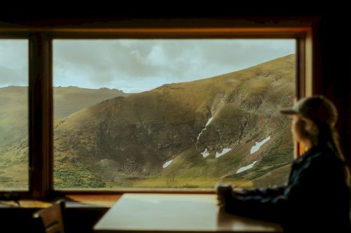 A person sits at a table by a window, gazing at a scenic mountain view with rolling hills and patches of snow.