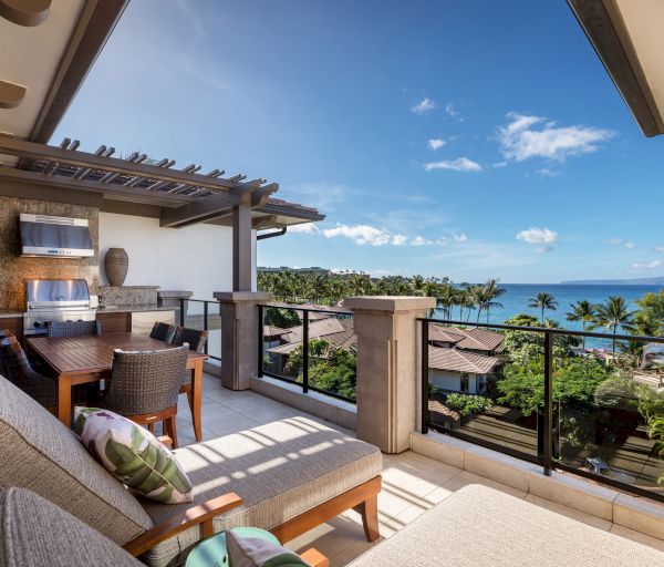 The image features a stylish balcony overlooking lush greenery and ocean views, with comfortable seating and a dining area.