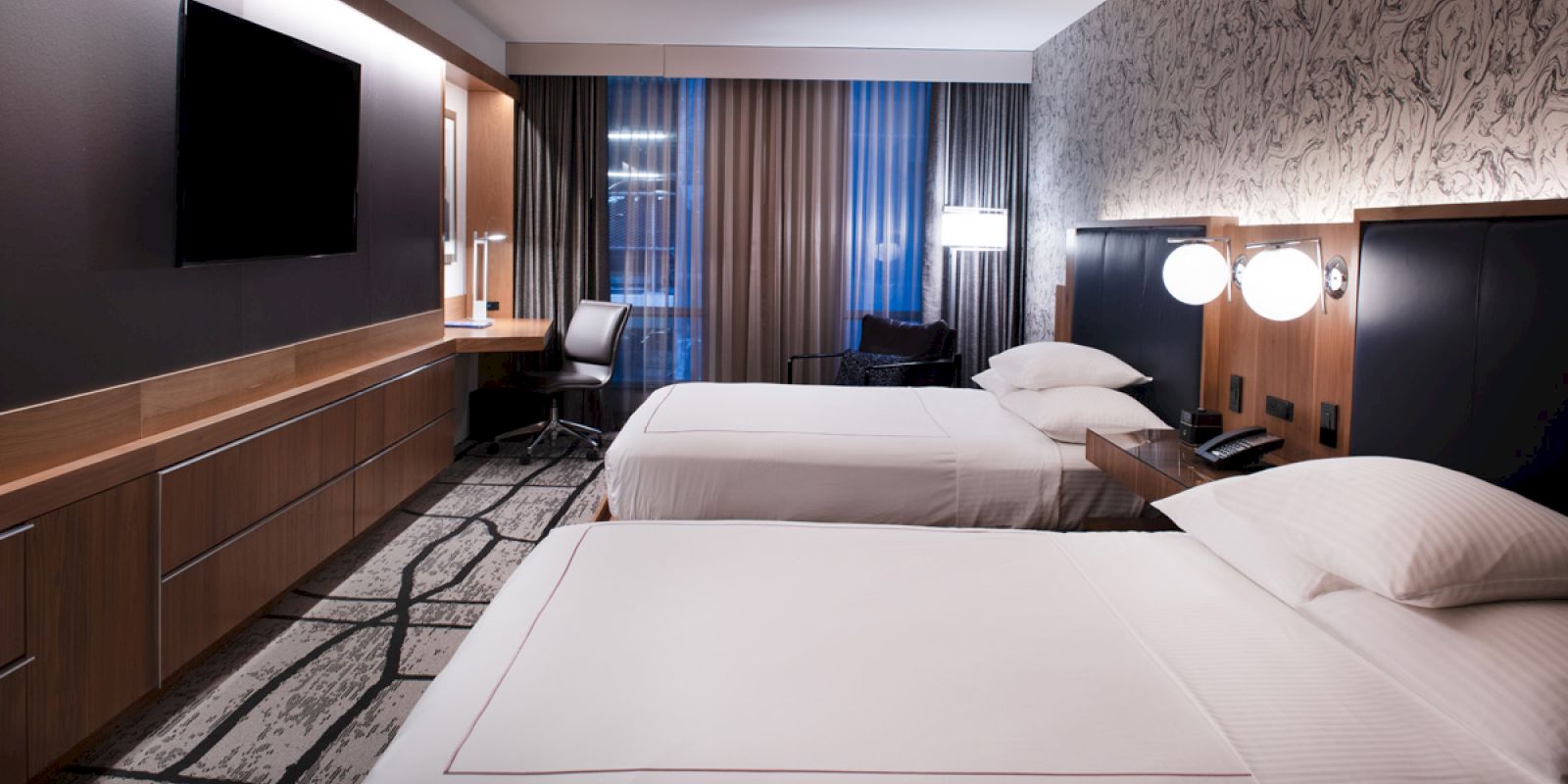 The image shows a modern hotel room featuring two beds, a desk, and contemporary decor. Soft lighting creates a cozy atmosphere.