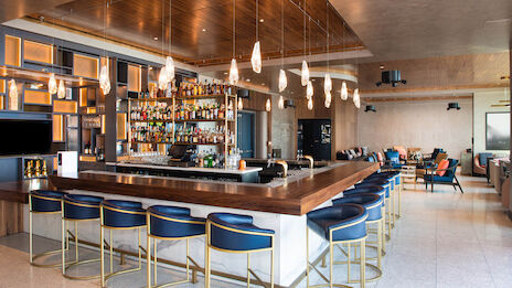 The image shows a stylish bar with a wooden counter, blue stools, and a well-stocked shelf of liquor bottles in a modern setting.
