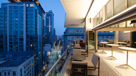 The image shows a sleek balcony overlooking a city skyline at dusk, featuring modern furniture and illuminated buildings.