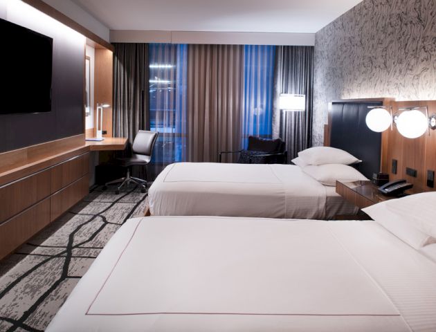 The image shows a modern hotel room featuring two beds, a desk, a TV, and stylish decor in shades of gray and wood.