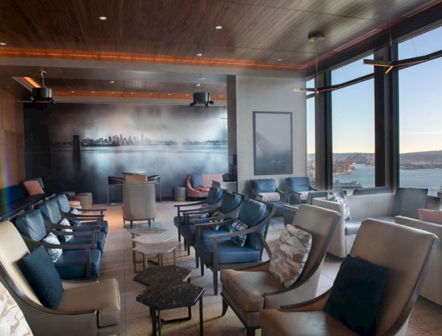 The image shows a stylish, modern lounge with comfortable seating, large windows, and a scenic view of the outdoors.
