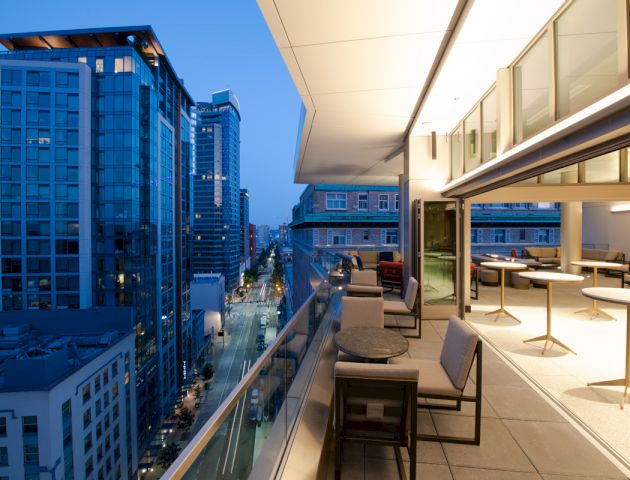 The image shows a modern balcony overlooking a city skyline at dusk, featuring sleek furniture and illuminated buildings.
