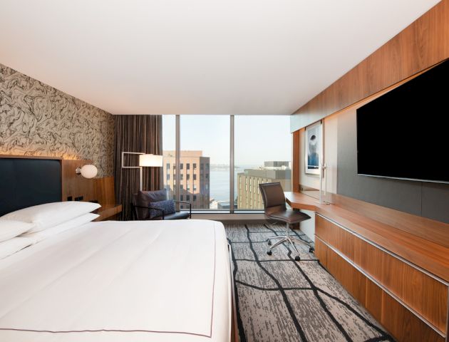The image shows a modern hotel room with a bed, a desk, a large TV, and a view through a floor-to-ceiling window.