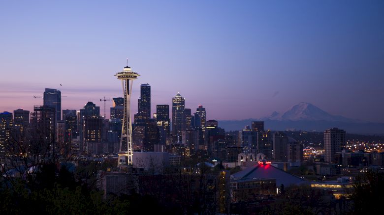 The image shows a city skyline at dusk with a prominent tower, likely the Space Needle, and distant mountains in the background.