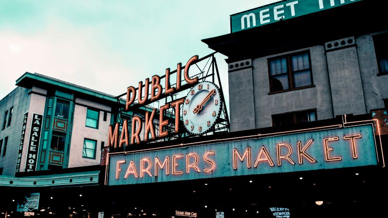 The image shows a neon sign for a public farmers market, with buildings in the background and an overcast sky.